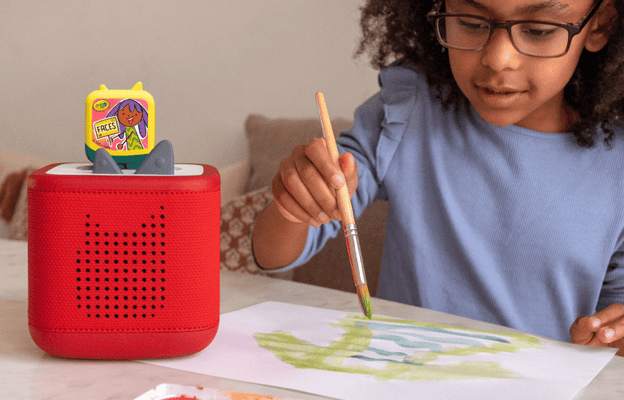 young child painting with watercolors, listening to a Crayola Art Adventures Pocket Tonie on a red Toniebox 2
